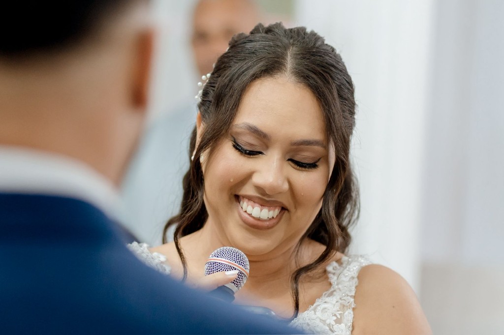 Noiva emocionada na cerimônia com vestido de renda, microfone e noivo ao lado em segundo plano.