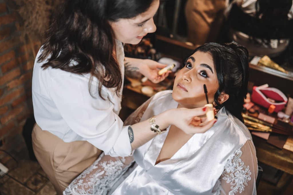 Maquiadora aplicando máscara de cílios em noiva de robe rendado, making of com penteadeira e espelho.