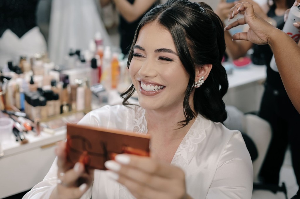 Noiva de robe branco sorrindo ao segurar paleta de maquiagem no making of, estação de beleza ao fundo.